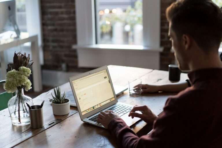 Man in een bordeaux trui typt op een laptop aan een houten tafel, met een vaas bloemen en een vetplant naast zich.