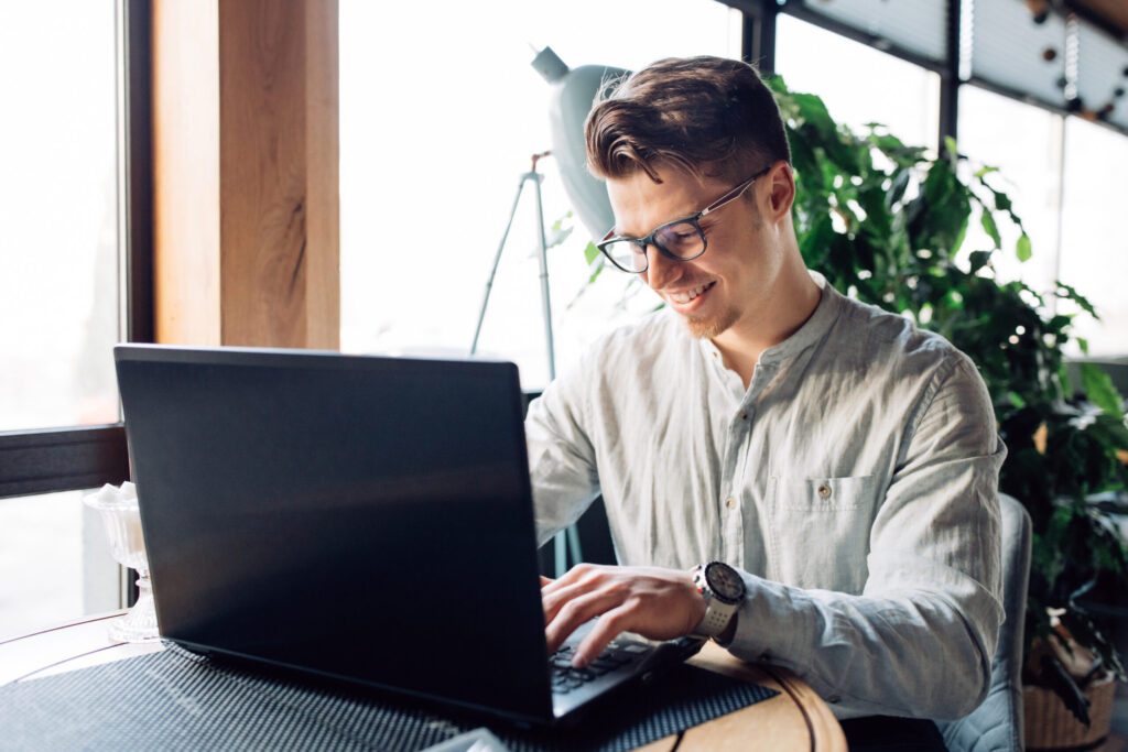 Man met een bril en een lichtblauw overhemd typt lachend op een zwarte laptop bij een raam.