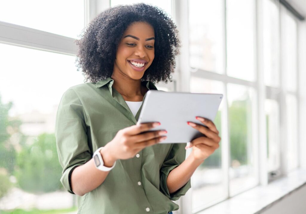 Vrouw in een groene blouse houdt een tablet vast en kijkt er lachend op, voor een rij hoge ramen.