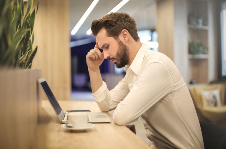 Man in een wit overhemd zit met zijn hand tegen zijn hoofd voor een laptop en een kopje koffie.