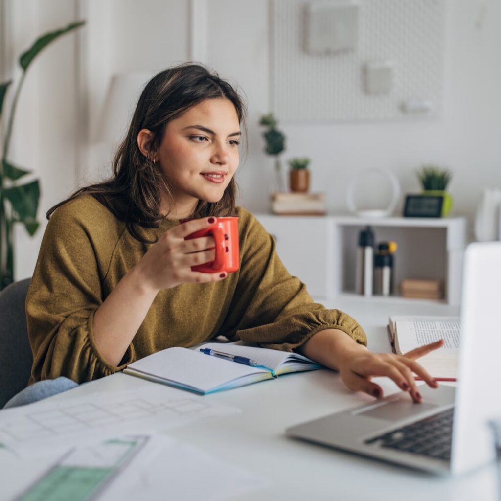 Vrouw aan een bureau met een laptop, een open notitieblok en bouwtekeningen, met een kop koffie in haar hand.