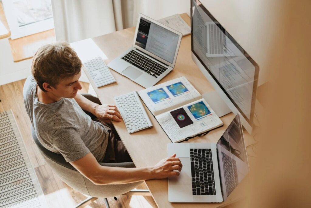 Man aan een houten bureau met twee laptops, een beeldscherm en een openliggend schrift met uitdraaien.