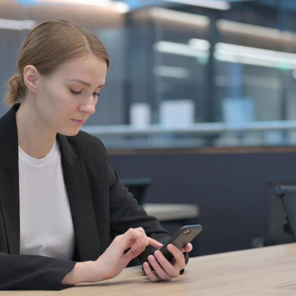 Vrouw in een colbert zit aan een lege tafel in een kantoor en kijkt op haar telefoon.