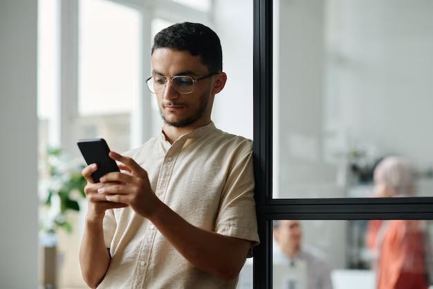 Man met een bril en een beige overhemd staat bij een glazen wand en leest iets op zijn telefoon.