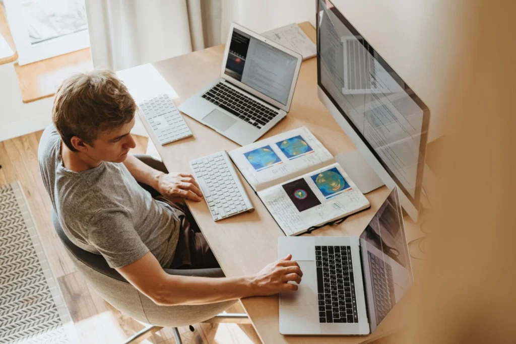 Man aan een bureau met twee laptops, een groot beeldscherm en een opengeslagen schrift